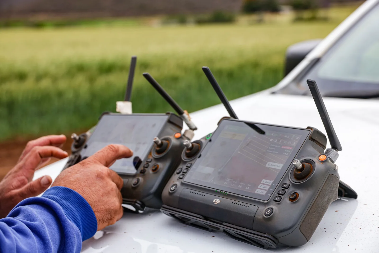Dos controladoras de drone apoyadas sobre el capot de una camioneta en el borde de un lote; una muestra en pantalla el mapa de aplicación con los límites del lote